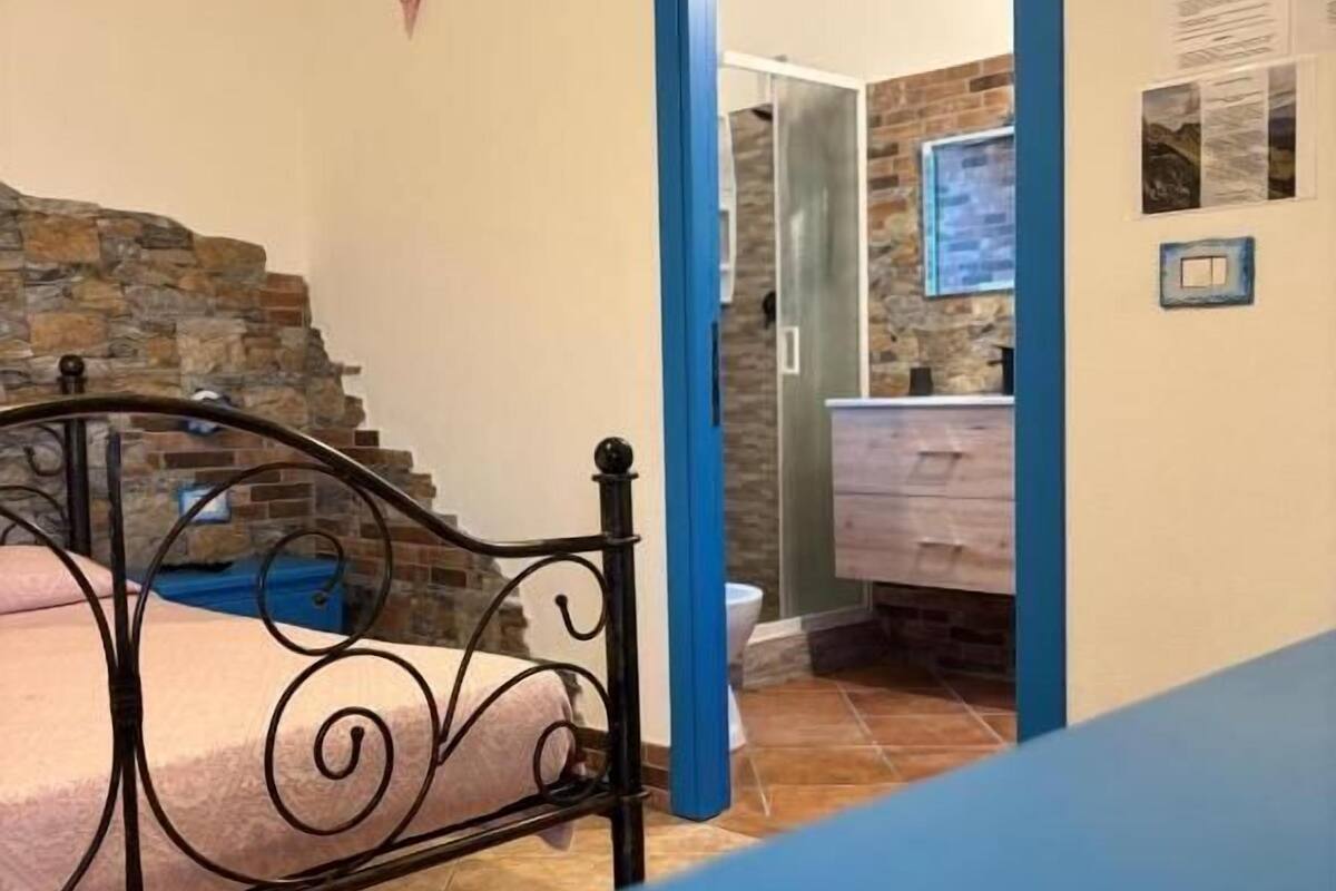 Un dormitorio con cama, una mesa azul, una pared de piedra y un baño visible a través de una puerta abierta.