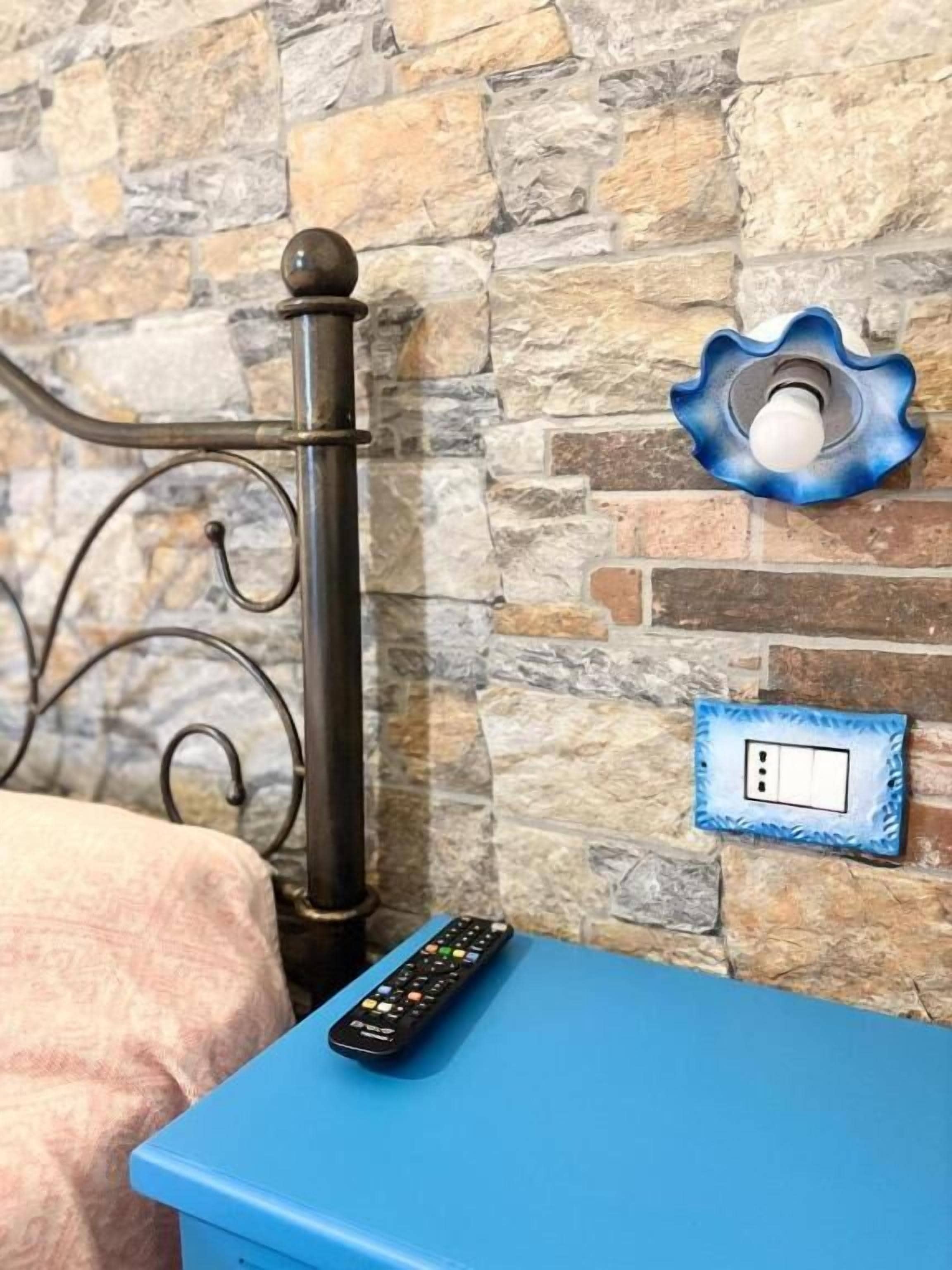 A bed with a metal headboard, a blue bedside table, a remote control, a wall-mounted light fixture, and a power outlet.