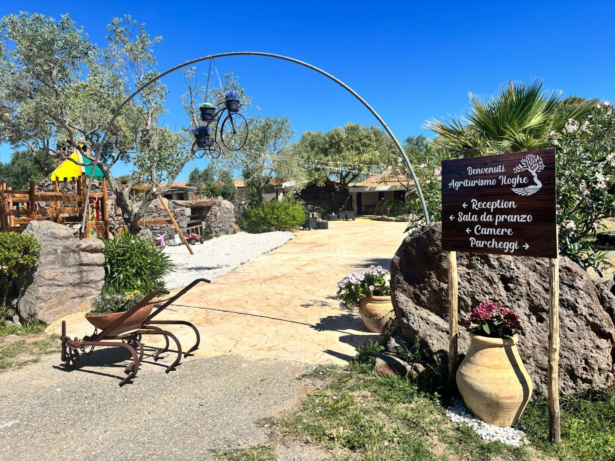 A rustic outdoor area with a wooden playhouse, a stone wall, and a sign indicating directions to reception, Sala da pranzo, and other facilities.