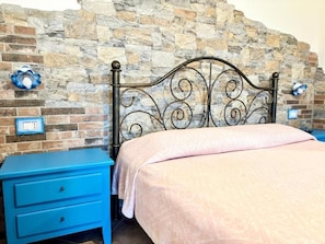 A bedroom with a stone wall, a metal headboard, a blue nightstand, and a pink bedspread. - Agriturismo Iloghe (Dorgali)