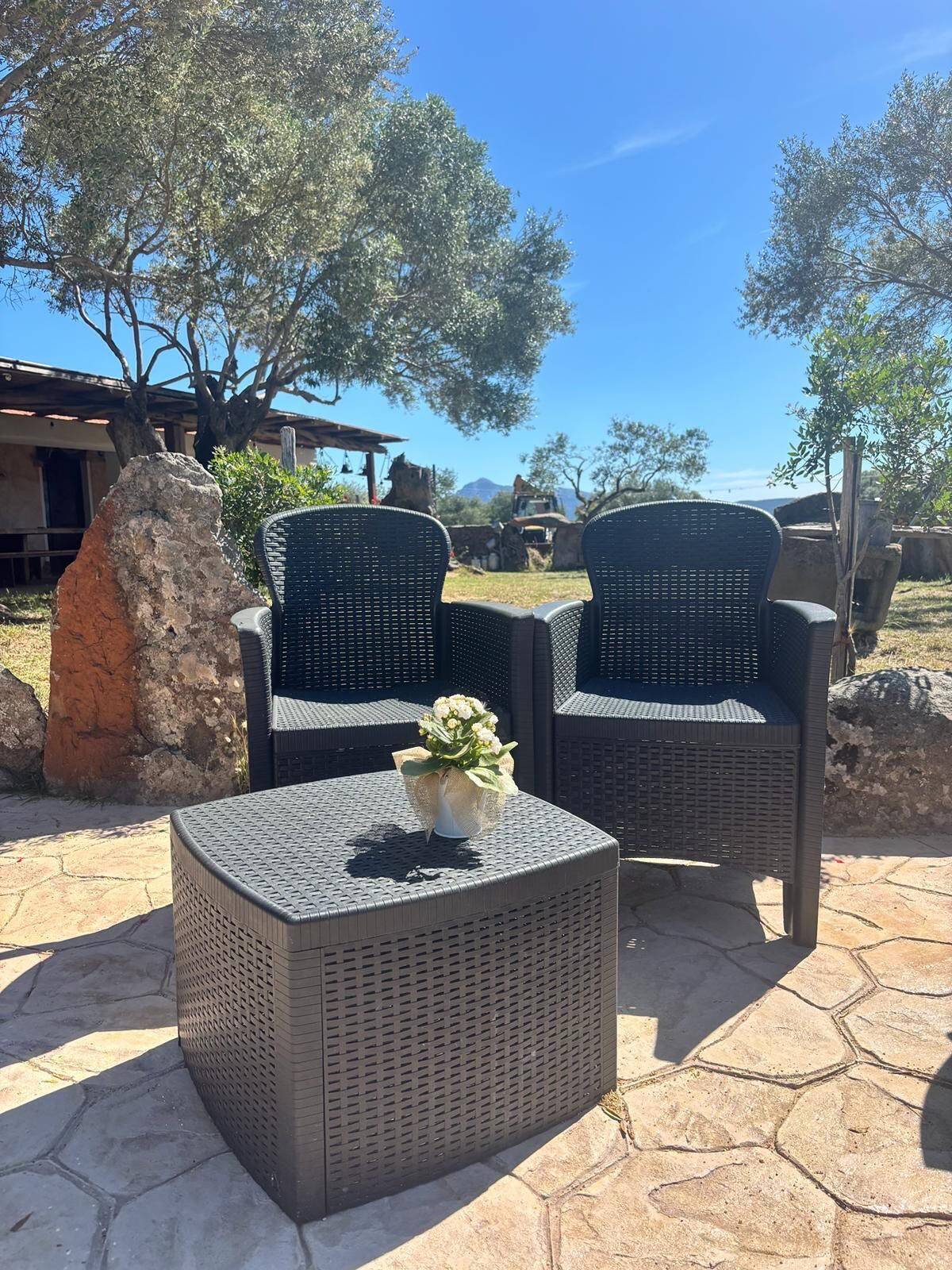 Mobiliário de vime ao ar livre com um arranjo de flores, colocado em um pátio de pedra com vista para árvores e um edifício ao fundo.