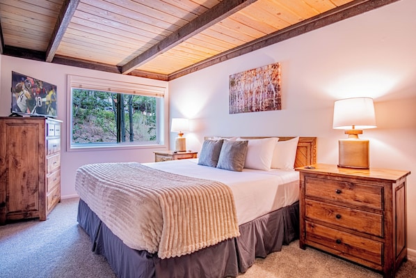 A bedroom with a large bed, two bedside tables with lamps, a wooden dresser, a window with a view of trees, and a painting on the wall. - Atelier Lookout - 2BD Condo Baldy Views + Pool (Sun Valley)