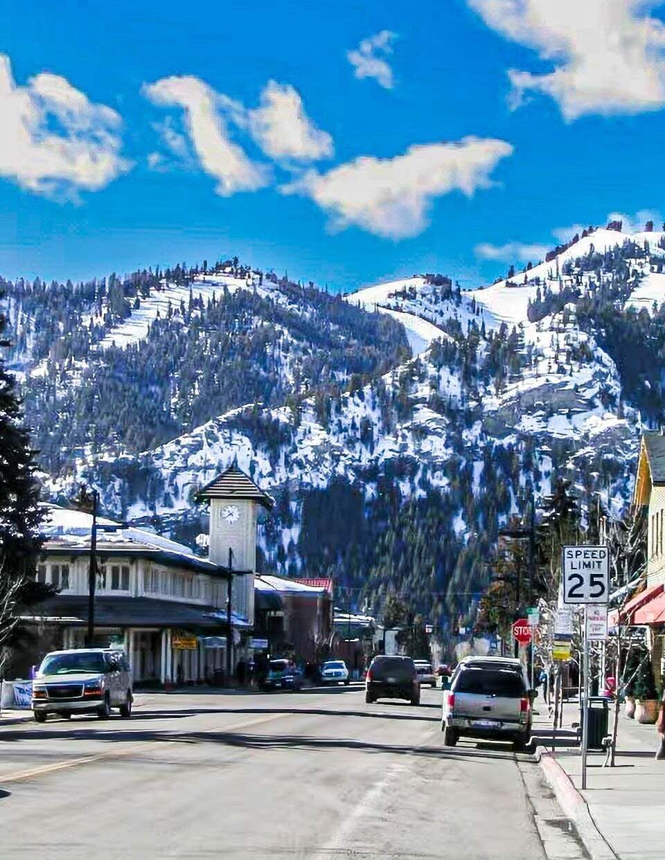 A town with a clock tower, a speed limit sign, and snow-covered mountains in the background.