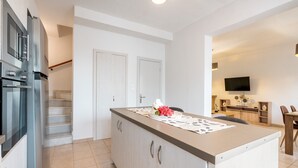 Una cocina moderna con una isla central, horno empotrado y zona de comedor con televisor y mobiliario de madera.