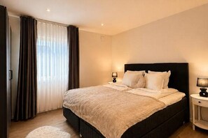 A bedroom with a large bed, two bedside tables with lamps, a window with curtains, and a door.