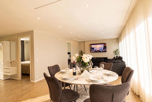 A modern dining area with a round table set for four, a sofa in the background, and a flat-screen TV mounted on the wall. - Seavista Trelleborg Sea View & Jacuzzi (Trelleborg)