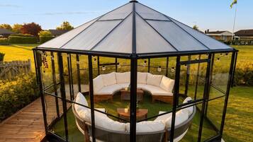 A glass gazebo with a curved white sofa and a wooden table, surrounded by a wooden deck.
