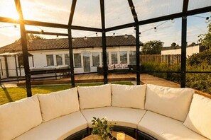 Un área de estar al aire libre techada con un sofá blanco curvo, una mesita con una vela y una planta, y vista a un edificio residencial al fondo.