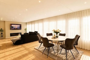 Una sala de estar moderna con chimenea, televisor montado en la pared, comedor con mesa redonda y sillas, y un ventanal con cortinas ligeras.
