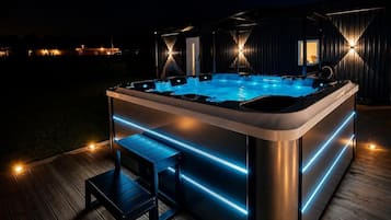Un jacuzzi con iluminación LED, una terraza de madera y un edificio moderno al fondo.