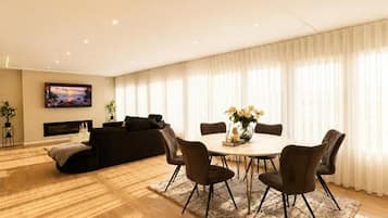 A modern living room with a fireplace, a TV mounted on the wall, a dining area with a round table and chairs, and a large window with sheer curtains.