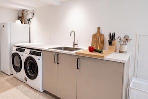 A compact kitchen with a washing machine, a sink, and a cutting board with vegetables. - Seavista Trelleborg Sea View & Jacuzzi (Trelleborg)