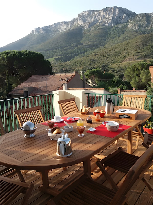 Une table en bois, dressée pour le petit-déjeuner, avec une vue sur les montagnes et une rambarde verte.