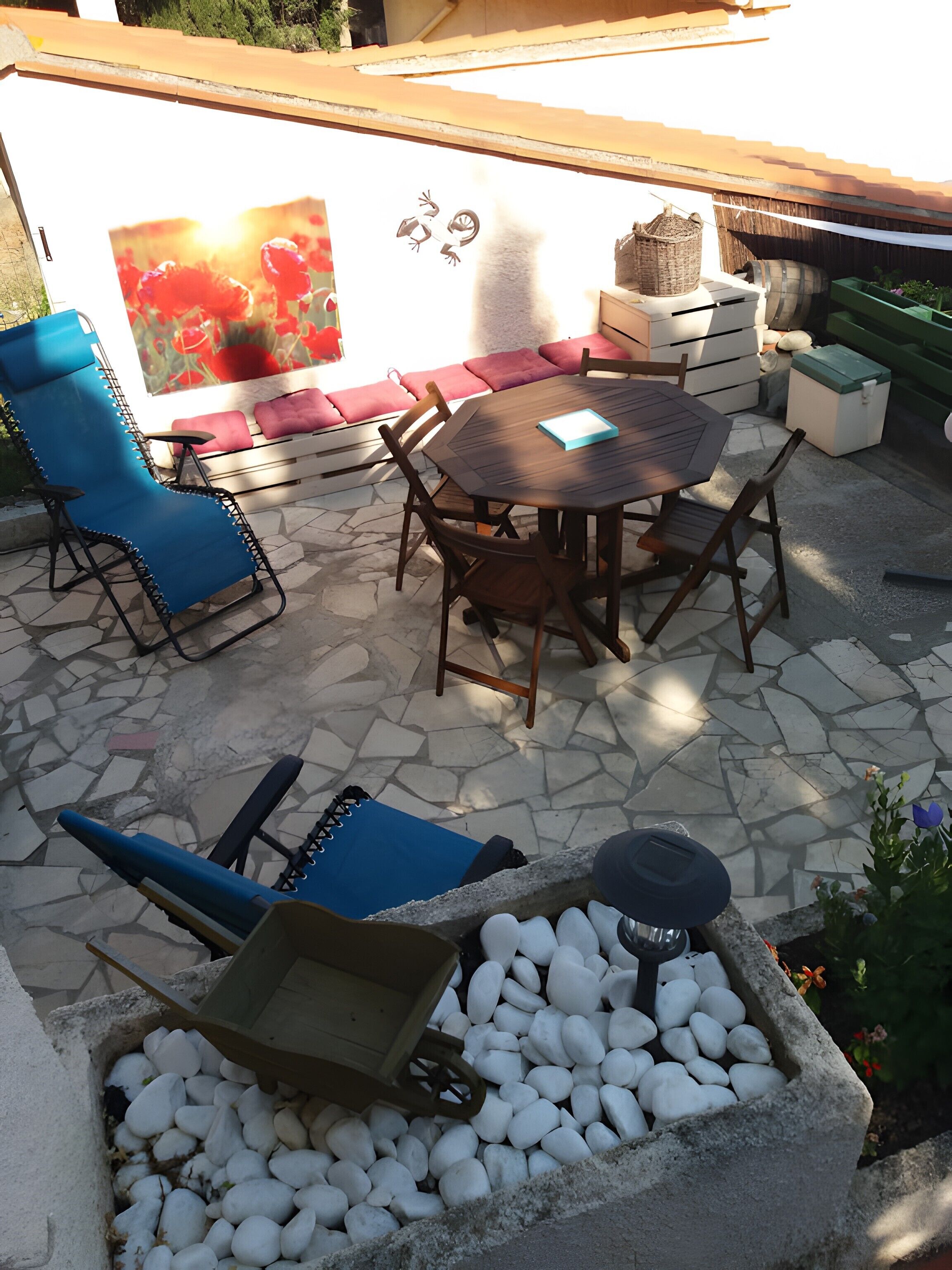 Un patio al aire libre con una mesa redonda, sillas azules, suelo de piedra y una pared con un cuadro.