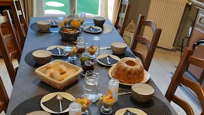 Ruokapöytä on katettu erilaisilla ruokalajeilla, siinä on hedelmäkulho ja kakku, ja pöydän ympärillä on tuoleja.
