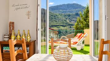 Un balcon donnant sur un paysage verdoyant et des montagnes, une table avec un vase, et un mur orné d’une inscription.