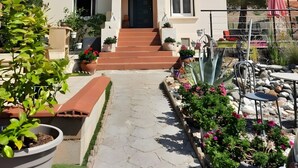 Un bâtiment à deux étages doté d’une entrée centrale, d’un balcon à l’étage supérieur et d’un jardin agrémenté de plantes en pot et d’une terrasse.