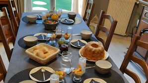 Une table à manger dressée avec différents plats, un saladier de fruits et un gâteau, entourée de chaises.