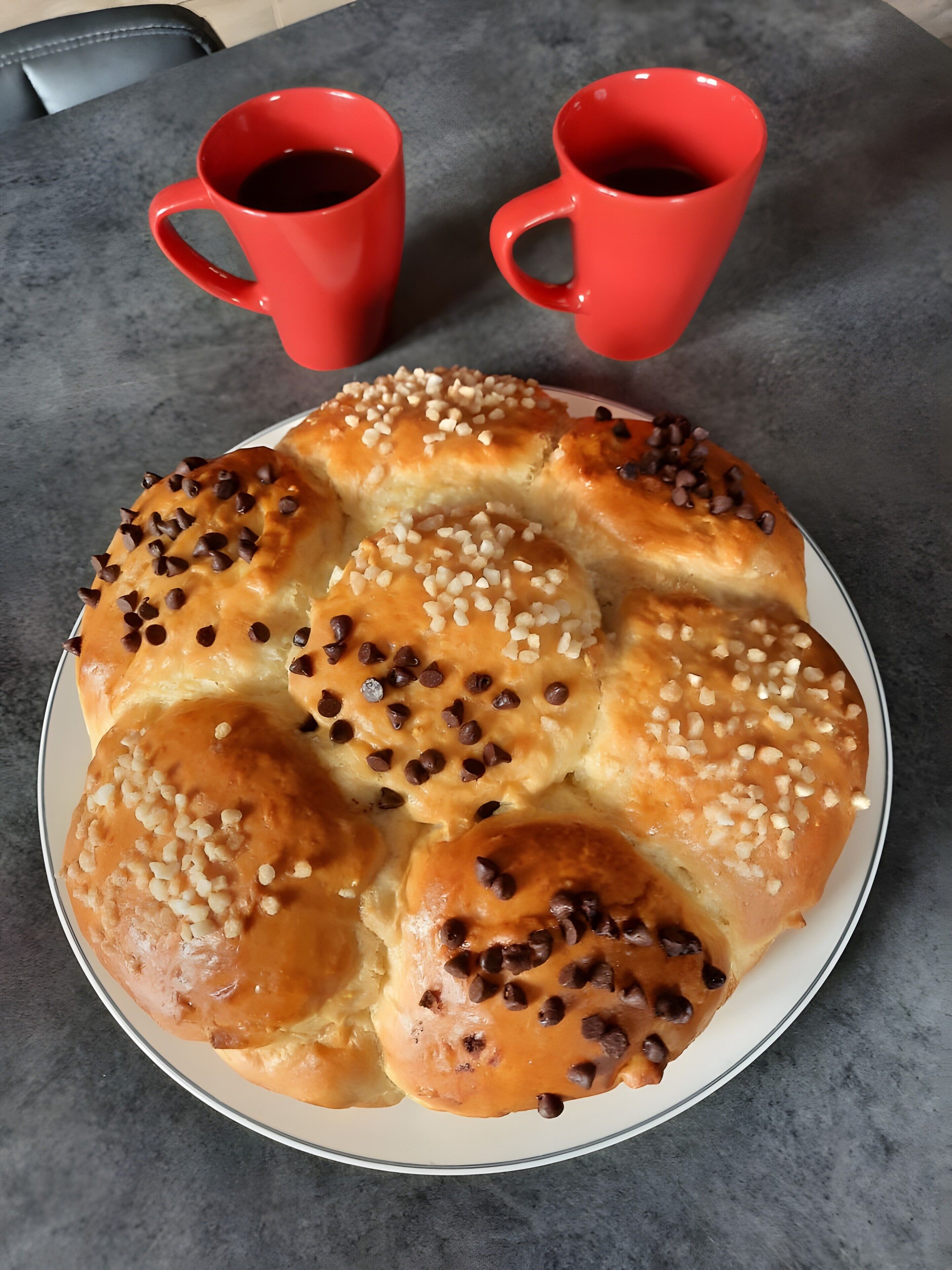 Een bord met broodjes met chocoladechips en noten, twee rode koffiekopjes, en een grijze tafel.