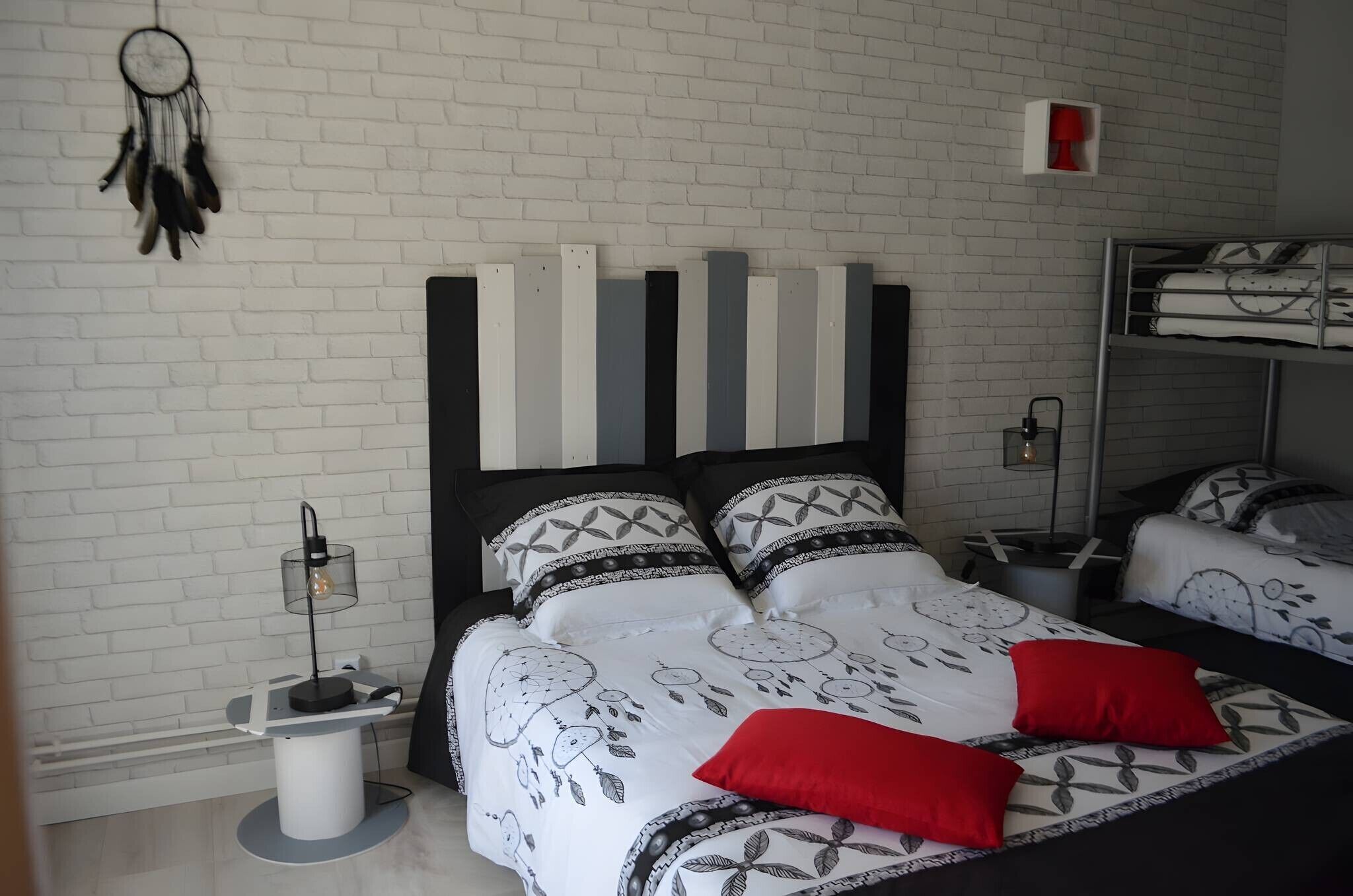 Um quarto com parede de tijolos brancos, uma cama com roupa de cama estampada, uma mesa de cabeceira com abajur e um apanhador de sonhos na parede.
