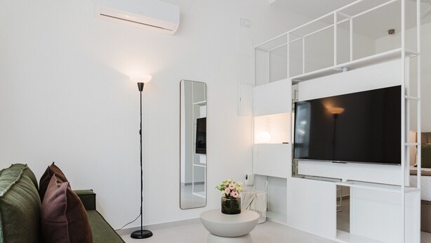 Casa de banho moderna com um espelho grande, sanita, lavatório e televisão montada numa parede branca.