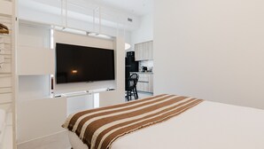 A modern bedroom with a bed, a TV mounted on a wall, and a kitchen area in the background. - Comfort and Design Facing the Turia Gardens in Valencia (Valencia)