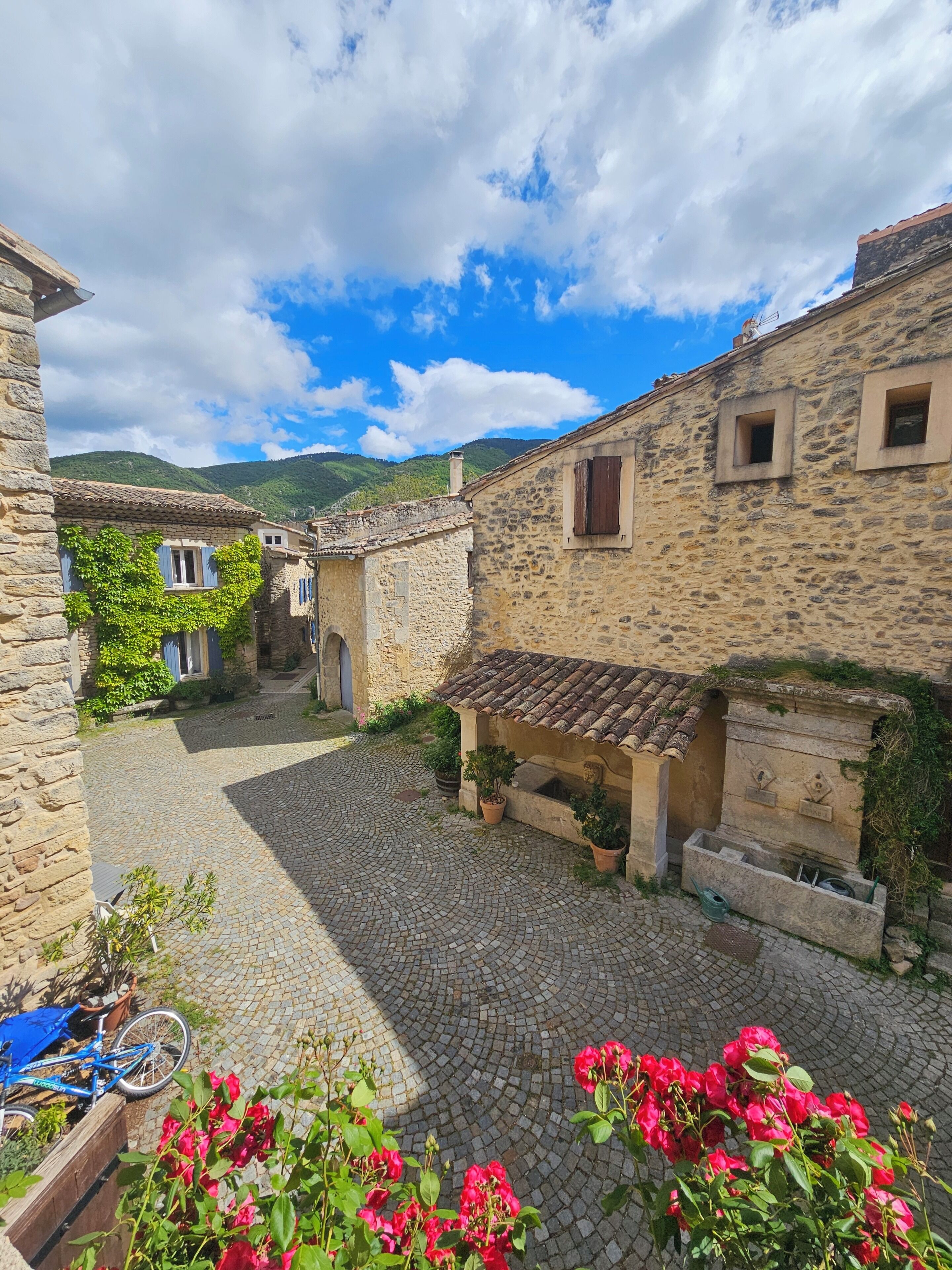 Une cour pavée de pierres, bordée de bâtiments en pierre, avec un vélo et des fleurs en pleine floraison.