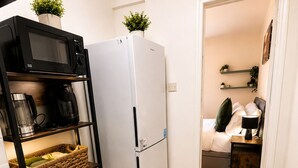 Cocina compacta con estufa, microondas y refrigerador. Se ven estantes con electrodomésticos y una cesta de mimbre. Una puerta da a un dormitorio con cama y mesita de noche.