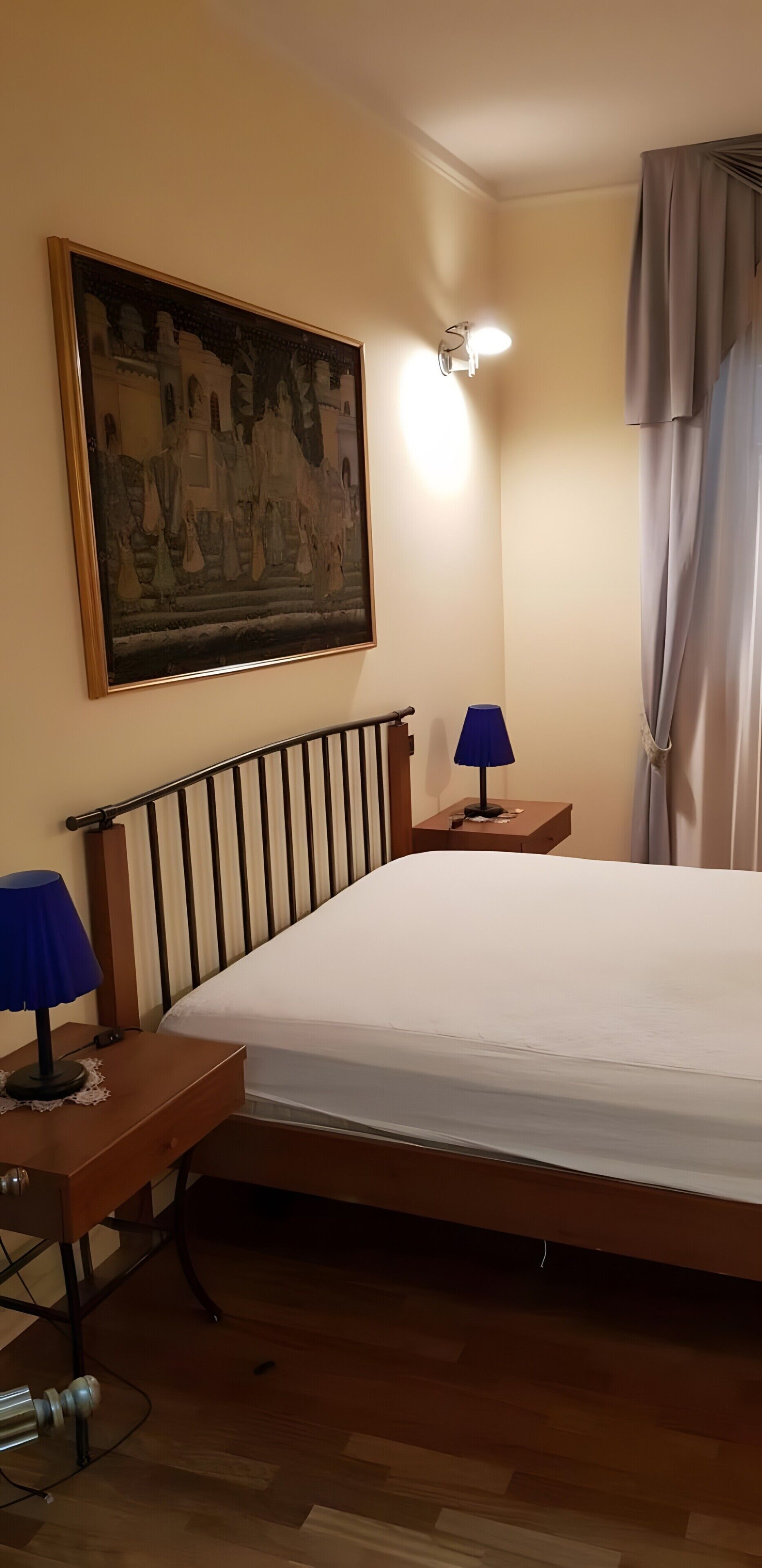 Una camera da letto con un letto, un comodino con una lampada, una luce a parete, un quadro e una tenda.