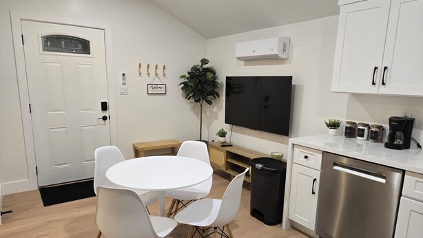 A modern kitchen with white cabinetry, a stainless steel dishwasher, a dining table with white chairs, a potted plant, and a flat-screen TV. - Cozy Guesthouse Near Disneyland in Walking Distance (Garden Grove)