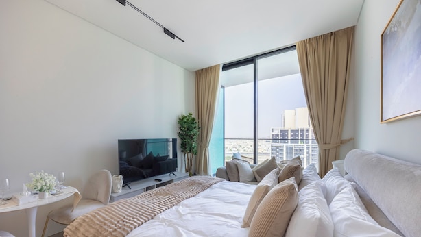 A modern hotel room with a large bed, a television, a desk with a chair, and a view of the cityscape. - Bloomfields Studio - Binghatti Azure JVC (Dubai)