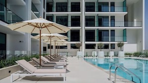 A pool area with lounge chairs and umbrellas, a swimming pool, and a modern building with balconies. - Bloomfields Studio - Binghatti Azure JVC (Dubai)