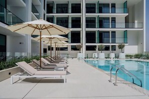 Uma área de piscina com espreguiçadeiras e guarda-sóis, uma piscina e um edifício moderno com varandas.
