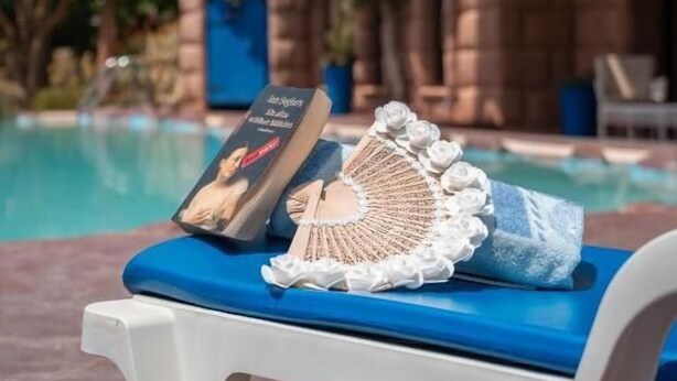 Una silla junto a la piscina con un libro y un objeto de crochet, un edificio que recuerda a un castillo al fondo, y un cielo azul despejado.