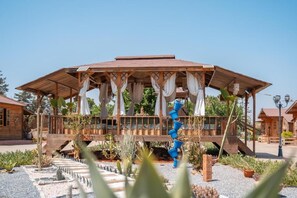 A wooden gazebo with a metal spiral sculpture, surrounded by a garden with various plants and a gravel path. - Lakala Complexe Touristique (Ben Slimane)