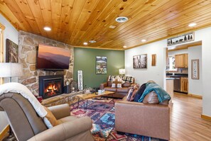 A cozy living room with a stone fireplace, a flat-screen TV, a comfortable sofa, and a kitchen in the background. - Webster Lodge at Covemont | Smoky Mountain Getaway (Sevierville)