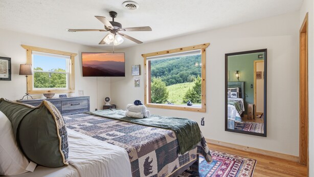 A bedroom with a bed, a ceiling fan, a TV, and large windows offering a view of a forest. - Webster Lodge at Covemont | Smoky Mountain Getaway (Sevierville)