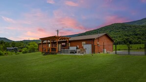 A wooden cabin with a porch and a covered outdoor area, surrounded by a well-maintained lawn and a forested hill in the background. - Webster Lodge at Covemont | Smoky Mountain Getaway (Sevierville)