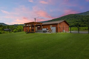 A wooden cabin with a porch and a covered outdoor area, surrounded by a well-maintained lawn and a forested hill in the background. - Webster Lodge at Covemont | Smoky Mountain Getaway (Sevierville)