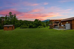 A wooden cabin with a porch and a small shed in a grassy field with trees and mountains in the background. - Webster Lodge at Covemont | Smoky Mountain Getaway (Sevierville)
