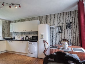 A compact kitchen with white cabinets, a washing machine, a microwave, and a dining table with a vase and placemats. - Keepers Cottage (Peterhead)
