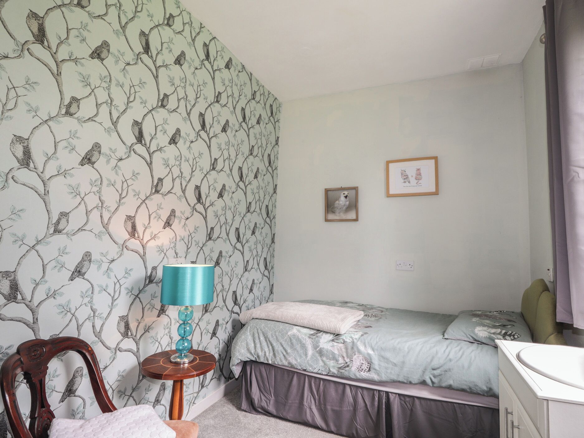 A bedroom with a patterned wallpaper, a bed with a grey and white duvet, a wooden chair, a small round table with a blue lamp, and two framed pictures on the wall.