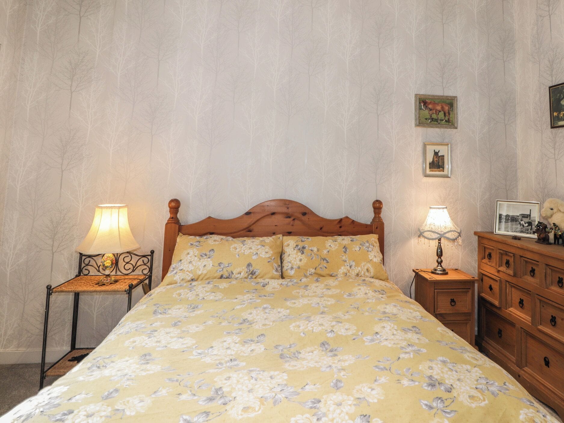 Un dormitorio con cama de madera, ropa de cama estampada, dos mesitas de noche con lámparas, un aparador y cuadros en la pared.