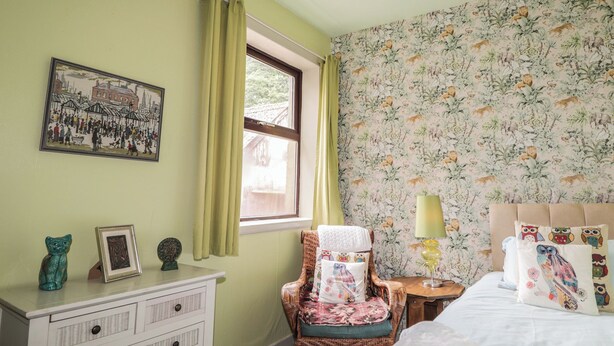 Quarto com cama, cadeira de vime, pequena mesa com abajur, cômoda e janela com cortinas.