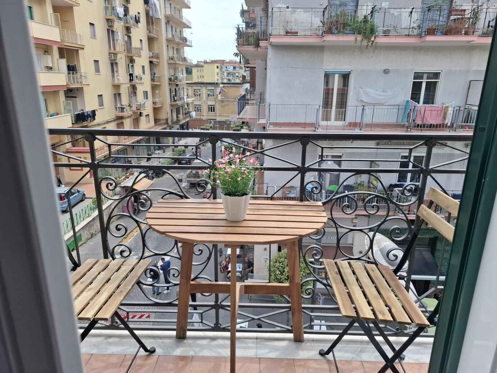 Un balcon avec une table et des chaises en bois, un pot de fleurs et une vue sur les bâtiments environnants.