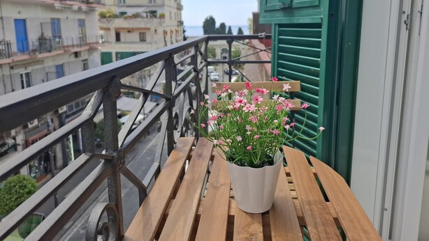 Ein Balkon mit Holztisch, einem Topf mit rosa Blüten und Blick auf Gebäude im Hintergrund.