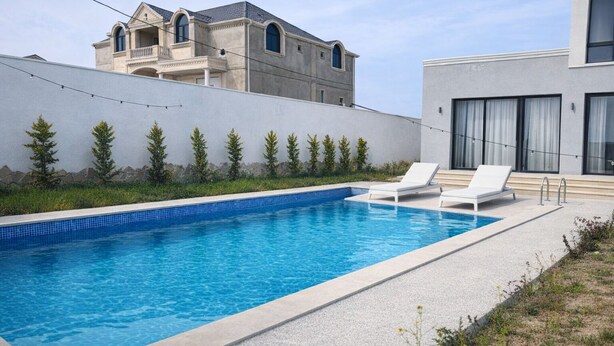 Una piscina rectangular con dos sillas reclinables blancas, un edificio moderno con balcón y un muro bajo con pequeños árboles.