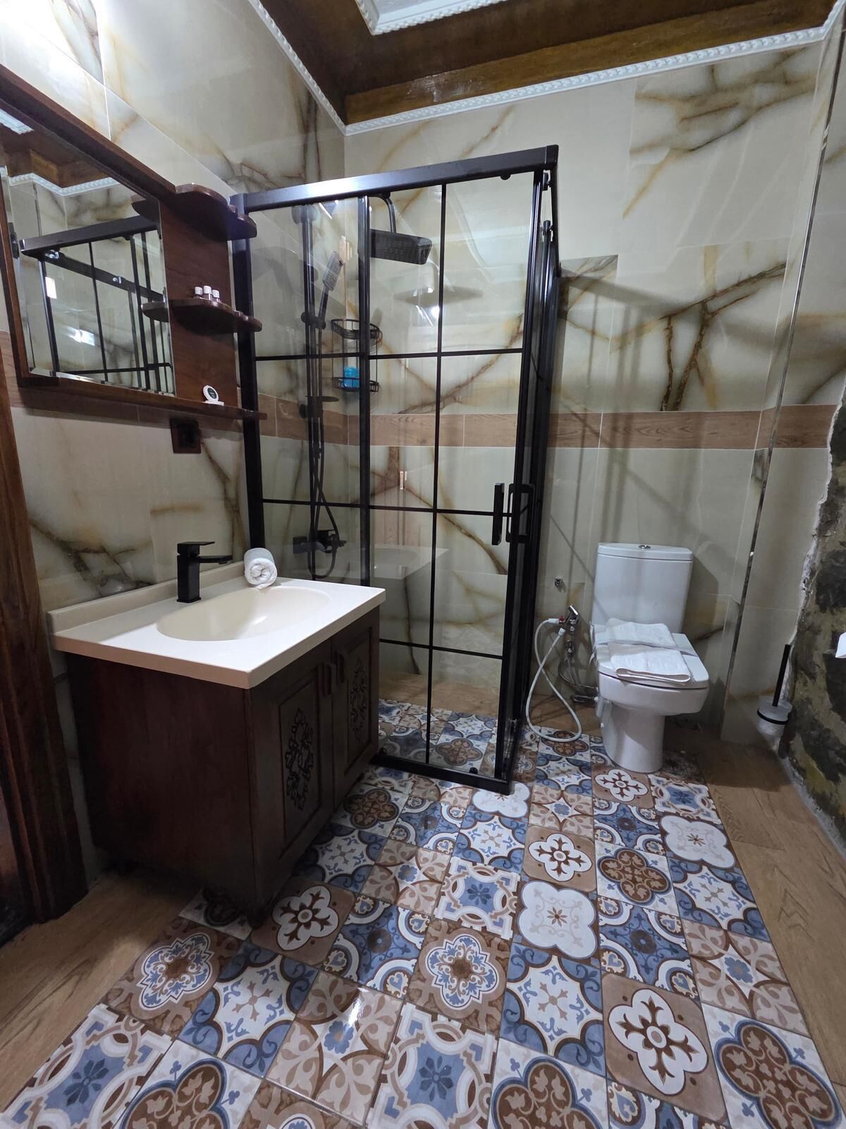 Un baño con pared de mármol, piso de baldosas, tocador de madera, cabina de ducha de vidrio y un inodoro.