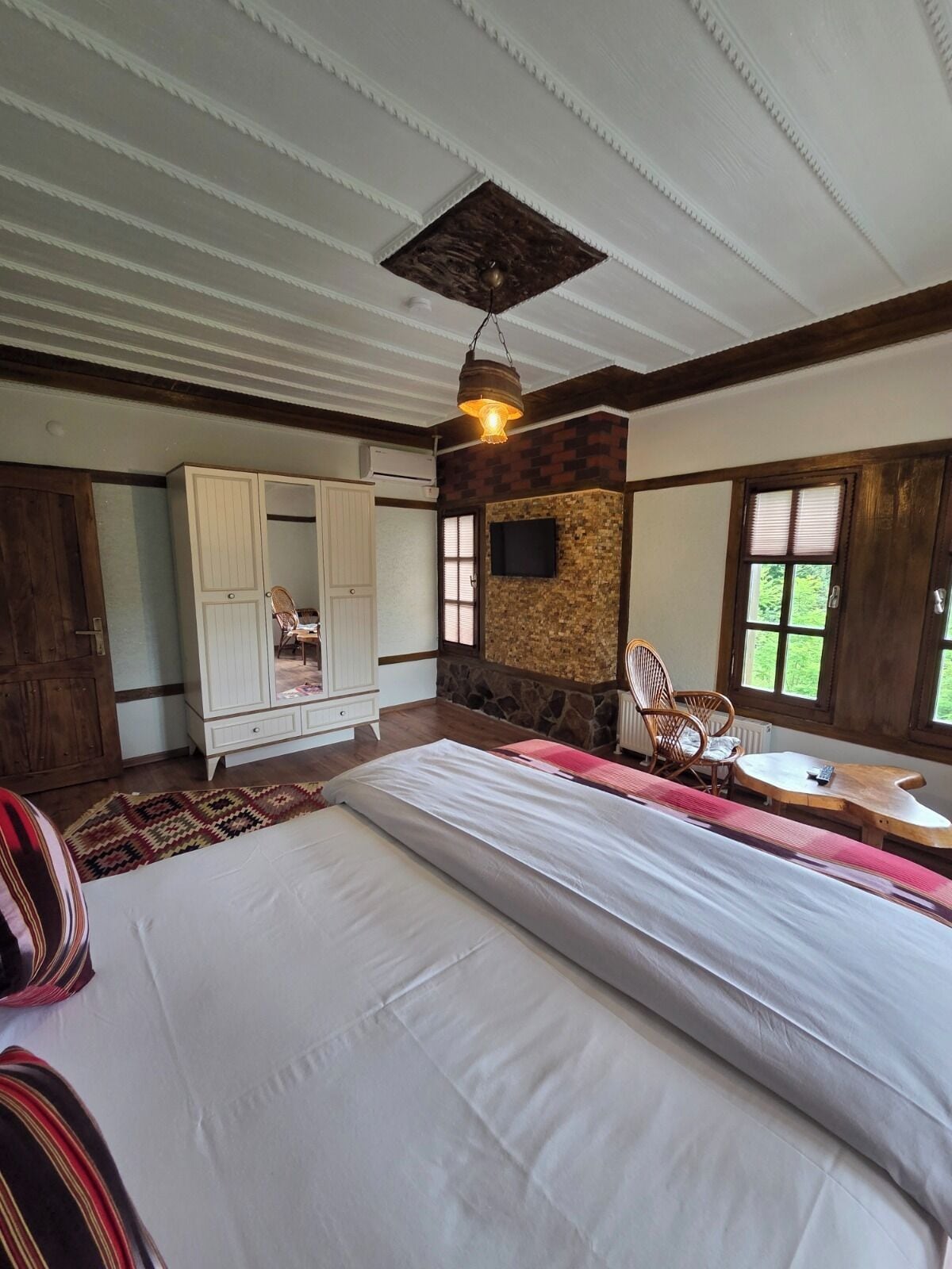 A bedroom with a large bed, a wardrobe, a stone accent wall, a television, and a window with blinds.