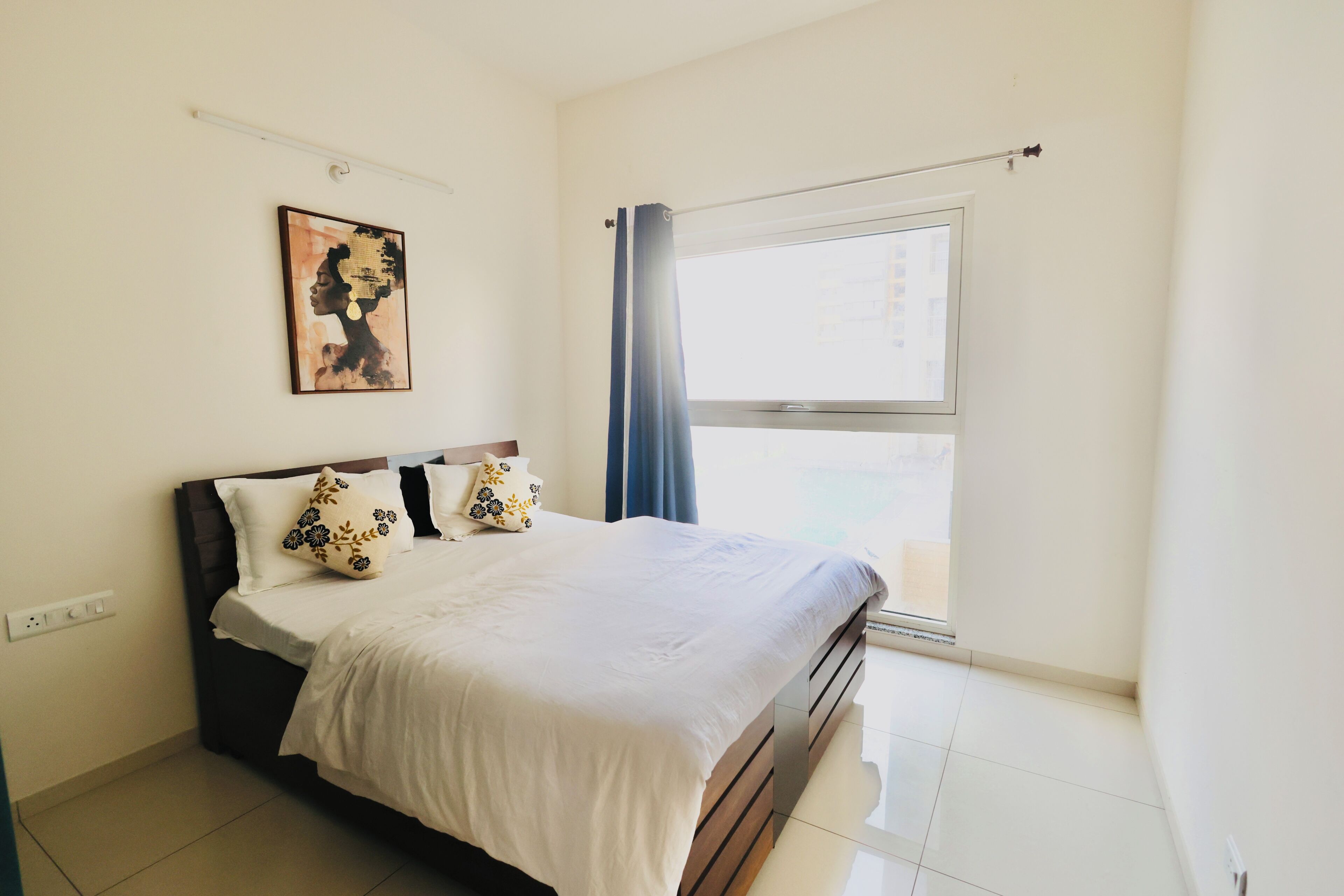 Un dormitorio con cama, ventana con cortinas, una obra de arte en la pared y un enchufe.